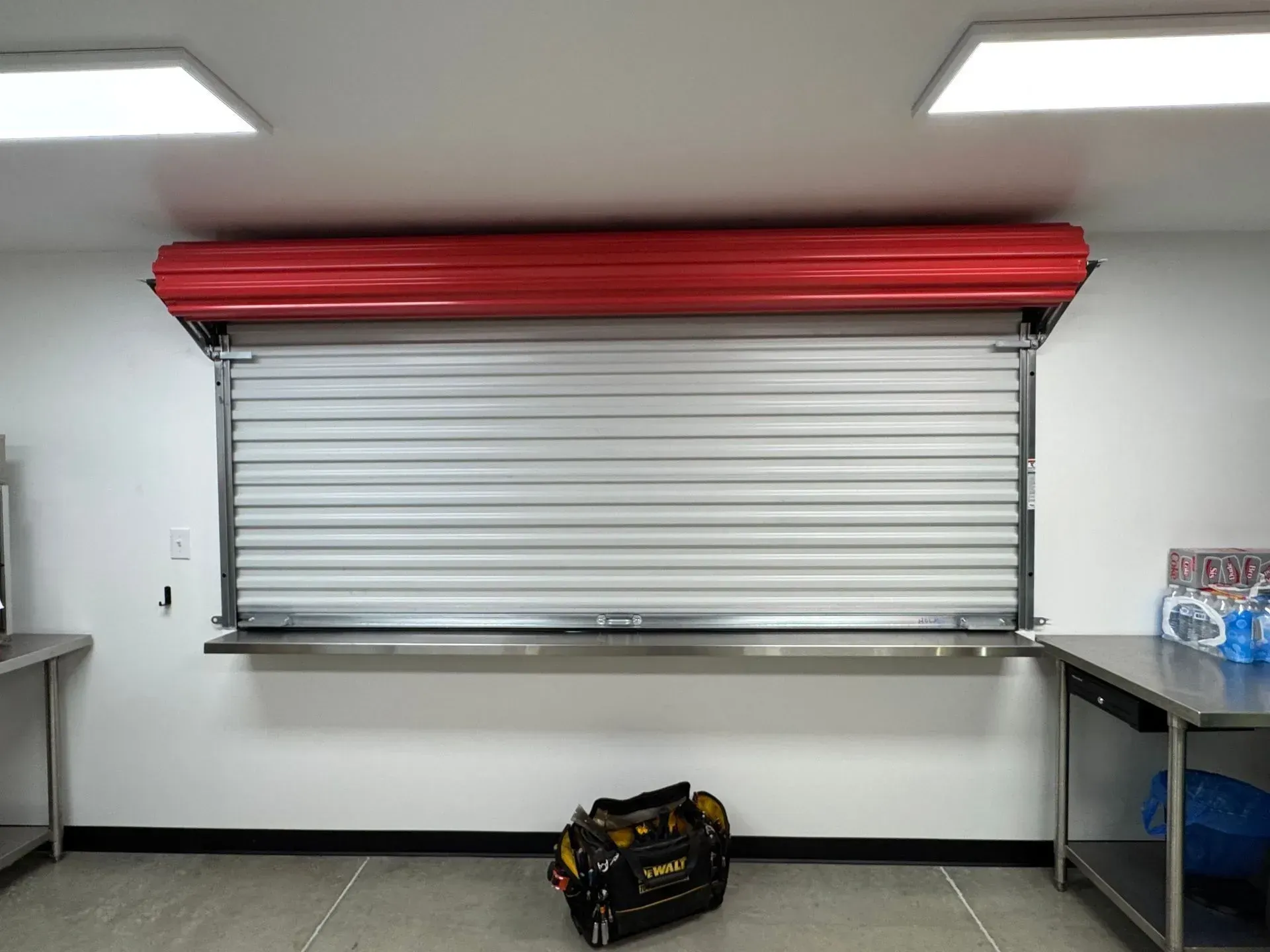 Closed white service window with red roll-up shutter in a bright room; tools bag on the floor below.