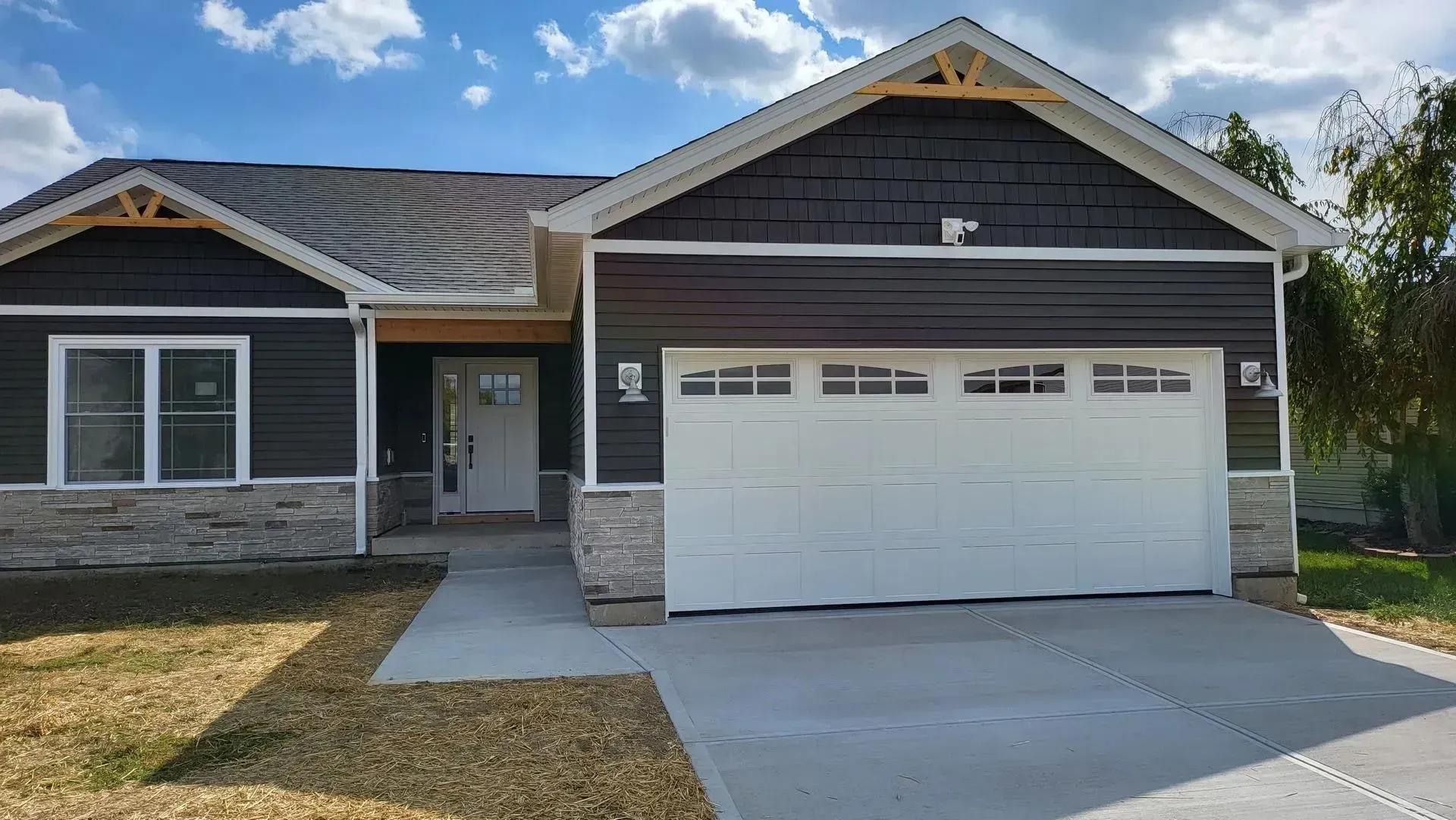 Suburban house with a white two-car garage, dark siding, and a concrete driveway under a blue sky