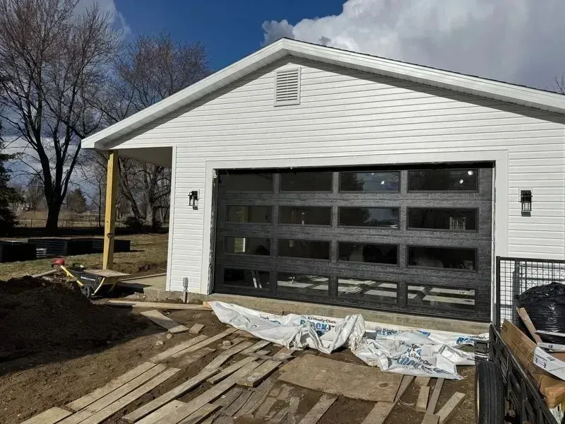 White garage with dark door under construction, surrounded by wood debris and dirt yard