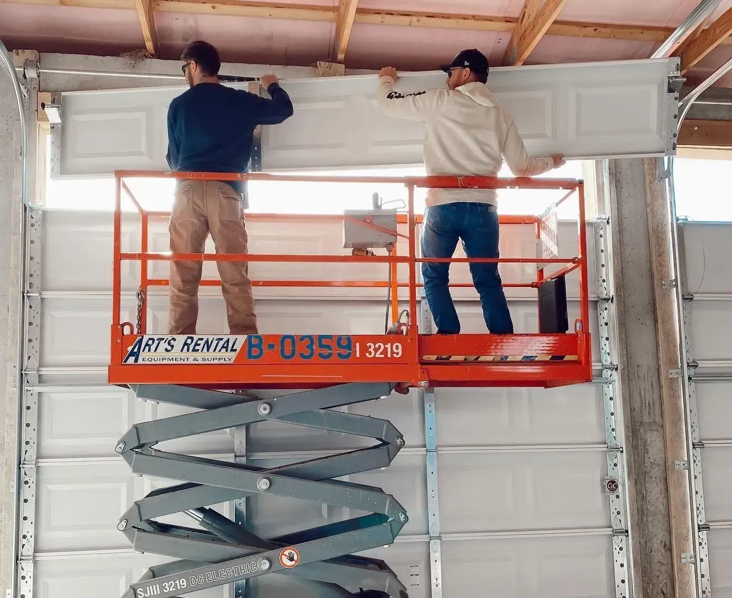 Two workers on an orange scissor lift installing a white garage door panel indoors