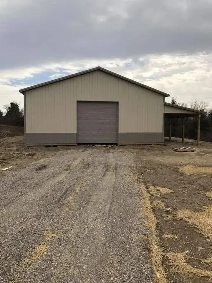 Gray metal barn with a large roll-up door on a gravel lot under cloudy skies