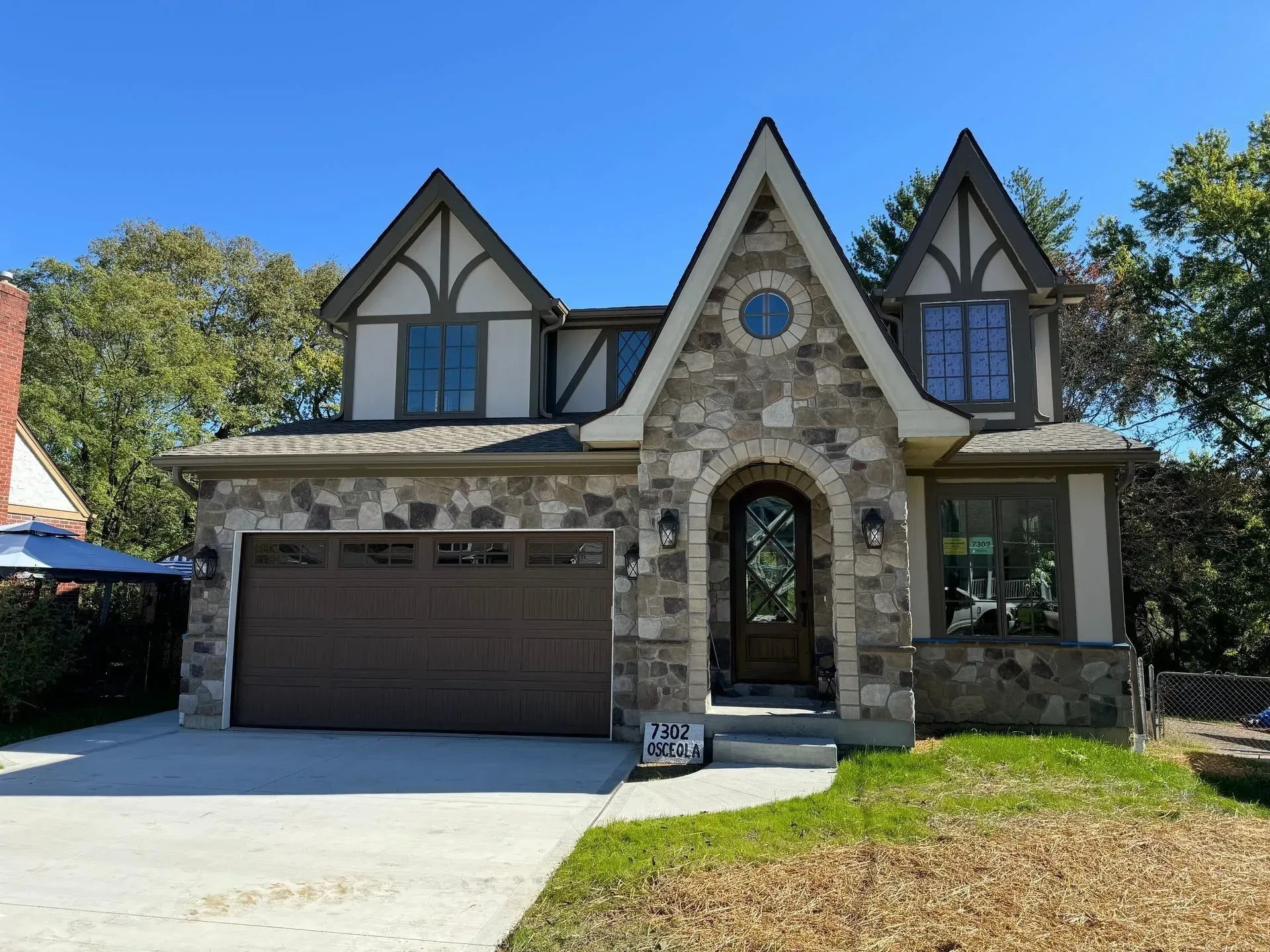 Stone Tudor-style house with a brown garage, arched stone entry, and gabled windows under a blue sky
