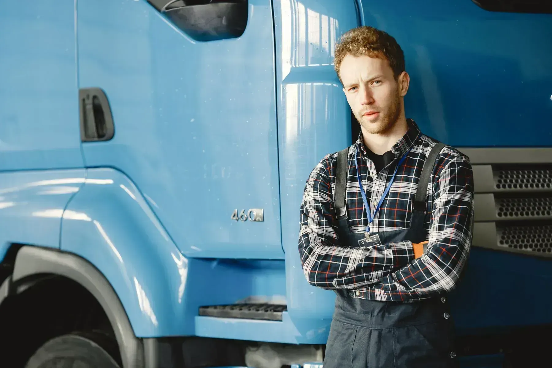 Man standing with arms crossed beside a blue truck, wearing a plaid shirt in an industrial setting