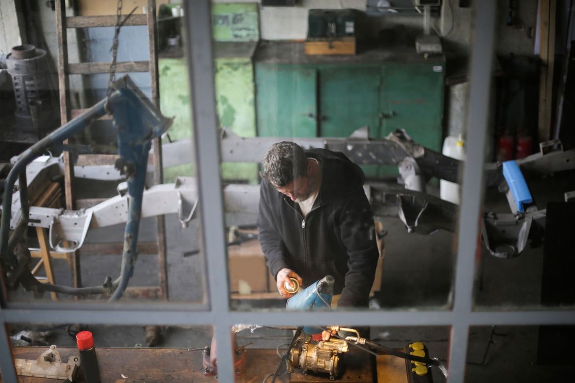 Person working with tools in a cluttered workshop, seen through a window.