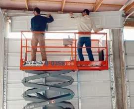 Two workers on an orange scissor lift installing a white garage door inside a building
