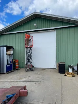 Industrial building exterior with a white roll-up door, lift equipment, and tools on the concrete apron.