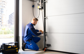 Technician kneeling beside a garage door opener, adjusting machinery in a bright workshop.