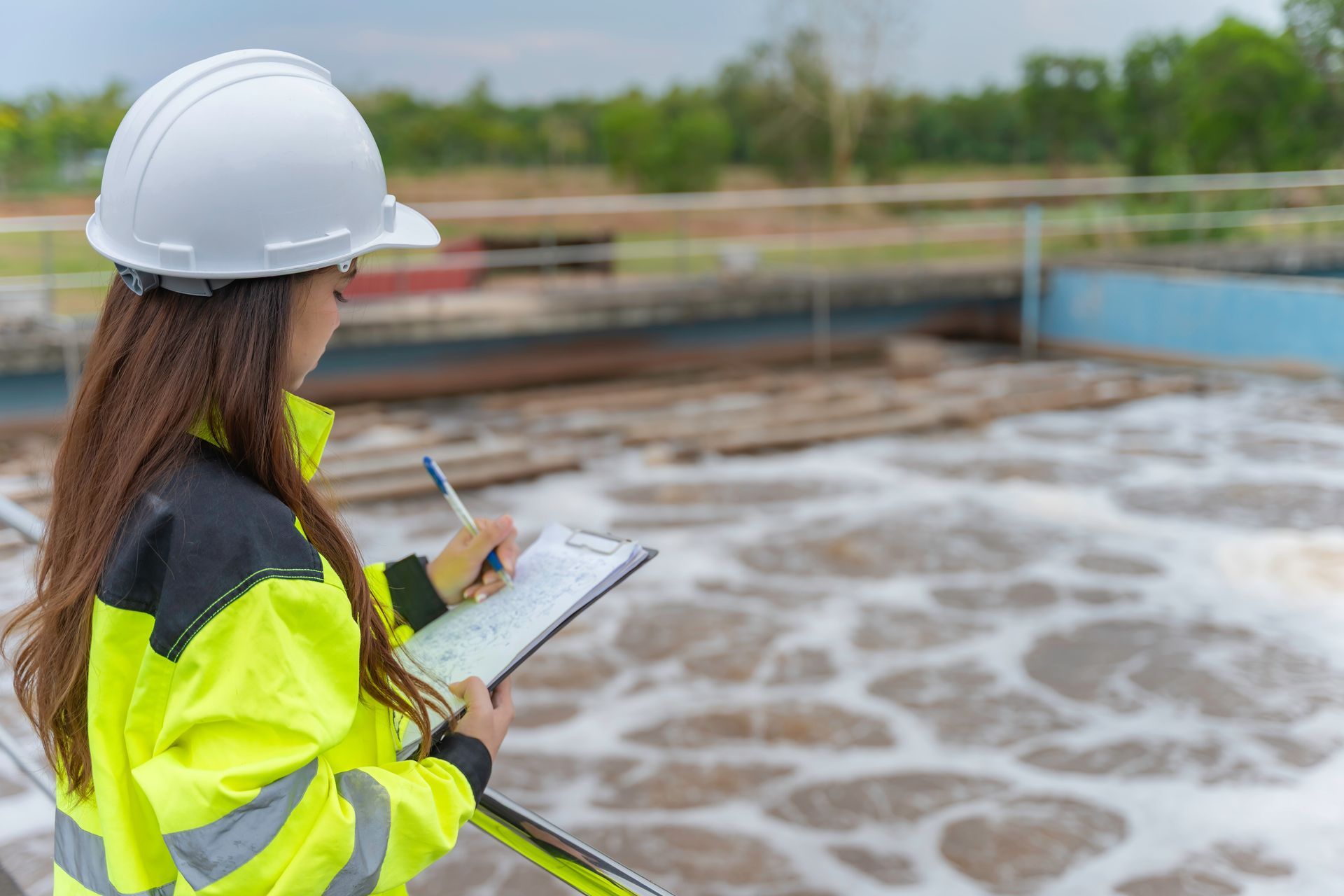 Worker in high-visibility jacket and hard hat inspecting water treatment plant with clipboard.