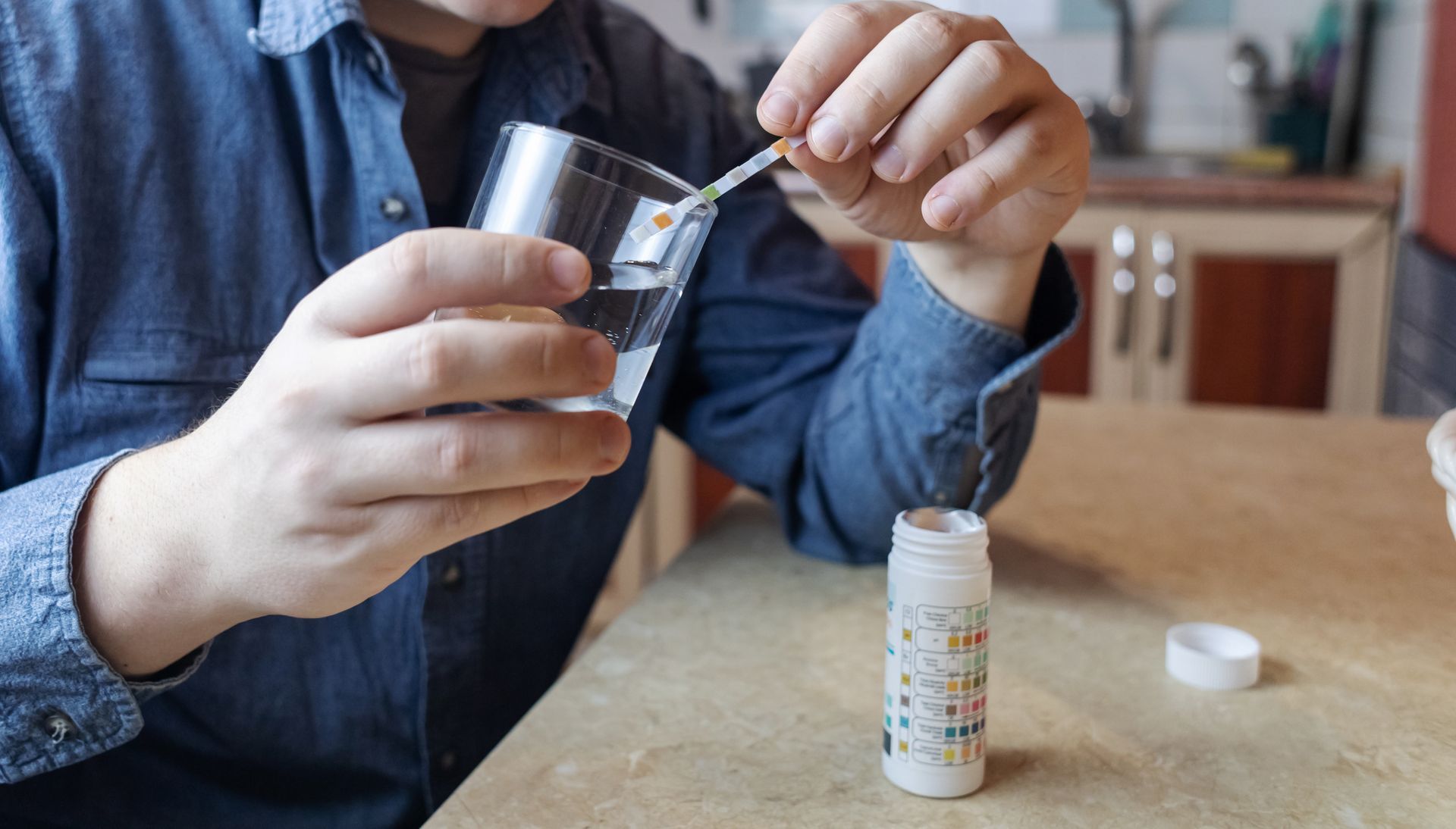A man in the kitchen tests tap water quality with a test strip, showcasing drinking water analysis. A man in the kitchen tests tap water quality with a test strip, showcasing drinking water analysis.