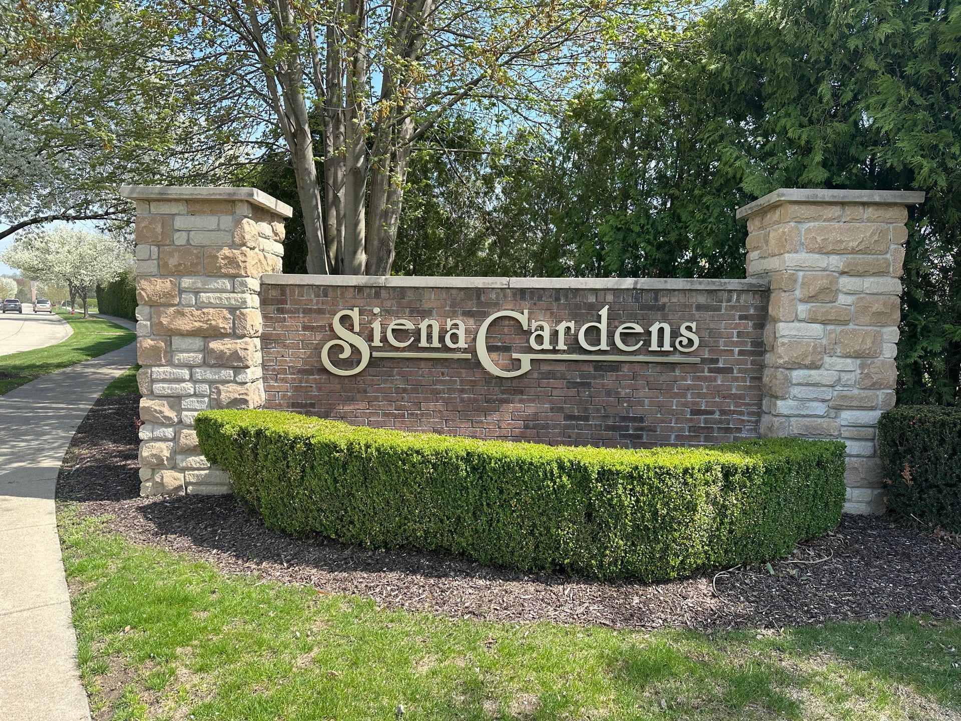 Stone entrance sign for Siena Gardens with brick pillars, trimmed hedge, and trees along a walkway.