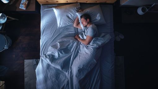 Person lying in bed, awake under gray sheets in a dimly lit bedroom.