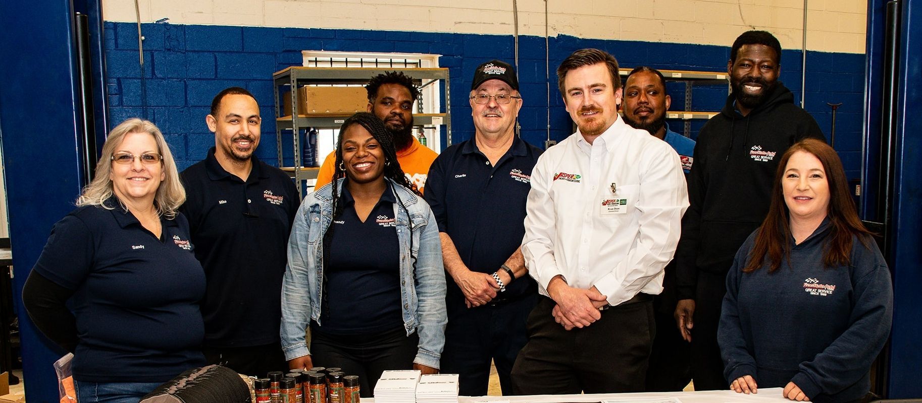 A group of people standing behind a table, smiling. They appear to be in a garage setting with blue walls | Freestate Auto & Truck Service