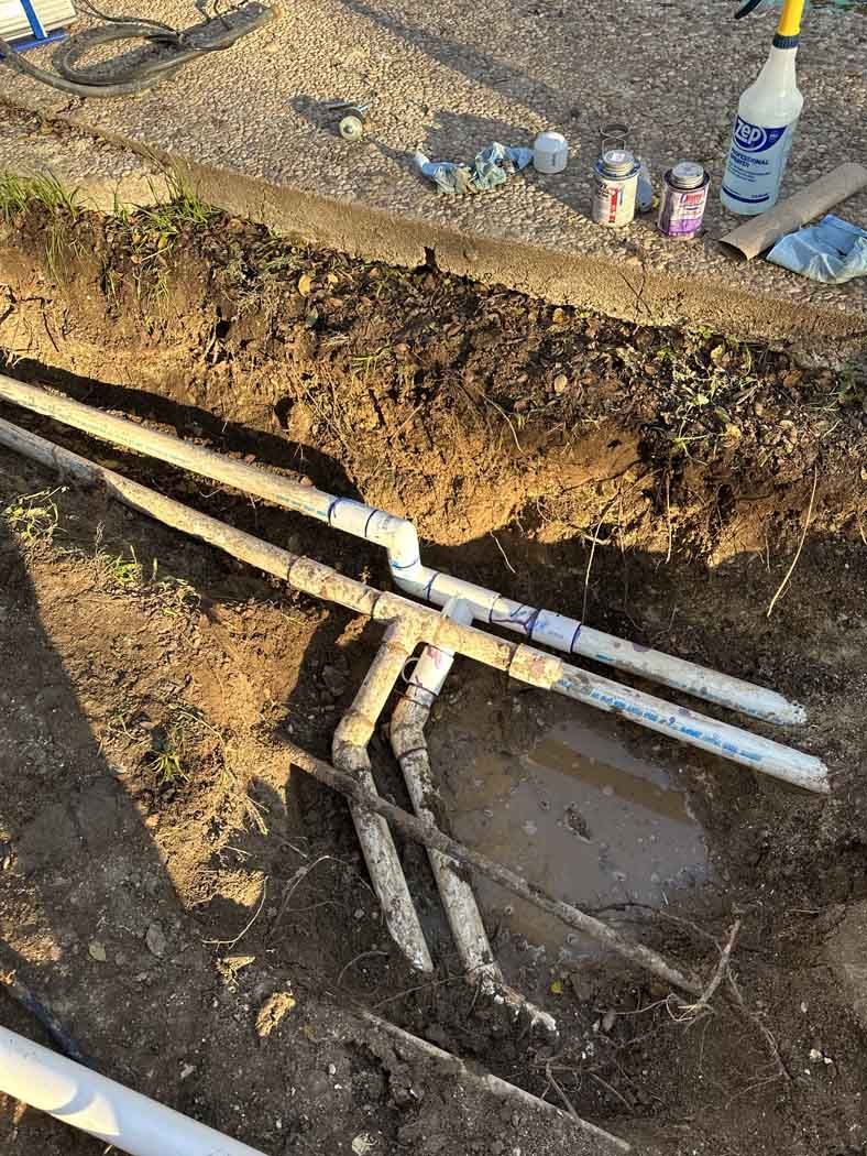 PVC pipes and fittings in a dirt trench, possibly for irrigation or plumbing.
