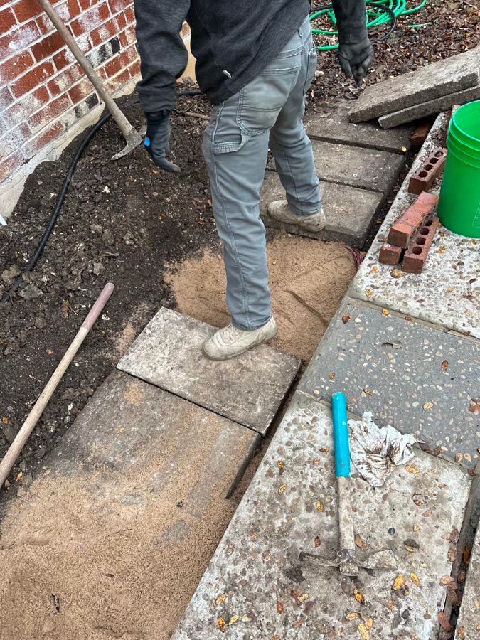 Person standing on concrete pavers, laying a walkway. Soil and sand are visible, with tools and bricks nearby.