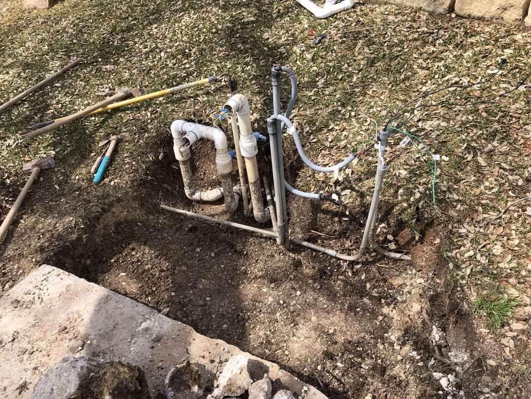 Exposed sprinkler system pipes in a dug-up patch of dirt on a lawn. Tools and debris scattered around.