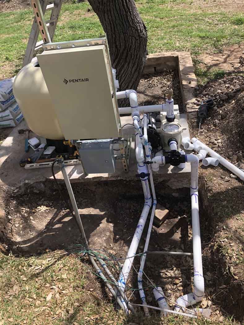 Pool equipment, including a filter and heater, sits near a tree in a yard. White PVC pipes connect the components.