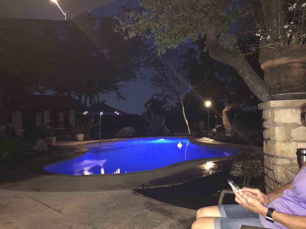 Nighttime view of a blue-lit swimming pool surrounded by trees, person using a phone on a patio.