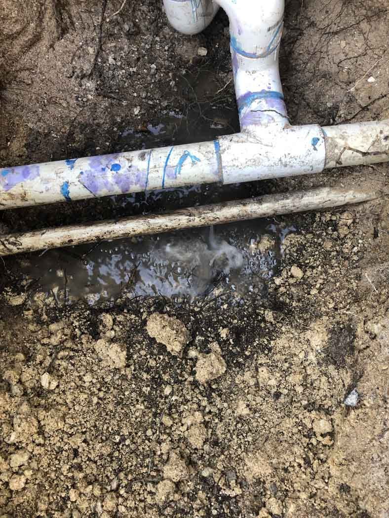 PVC pipe in a trench leaking water; white and purple pipe fittings against dirt.