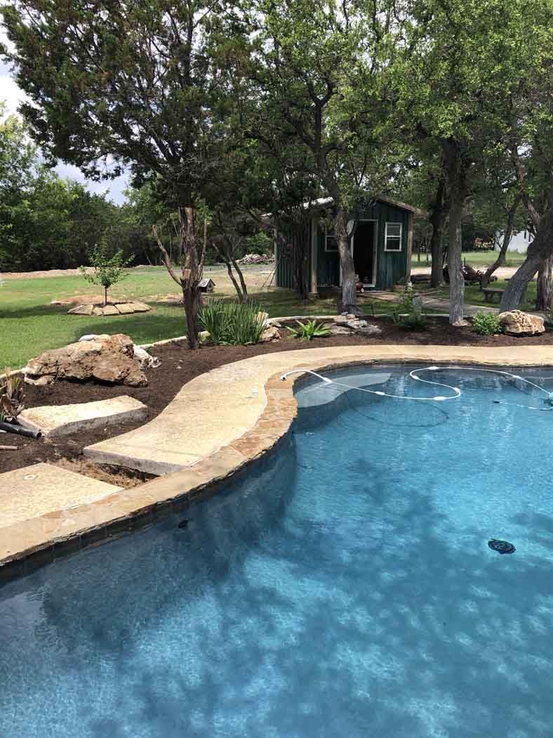 Pool with blue water and stone edge, concrete path, small green shed in background. Trees surround.