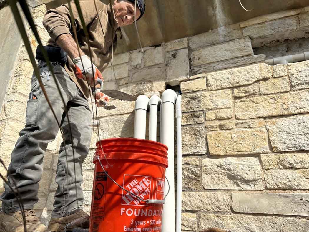 Construction worker using a trowel to work on stone wall near a bucket and pipes.