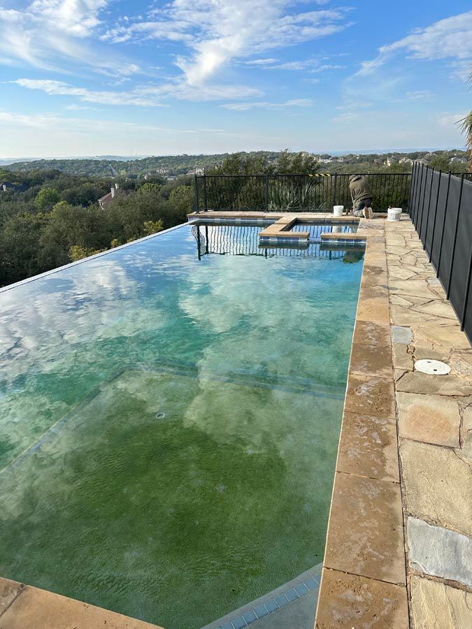 An infinity pool reflecting the sky, surrounded by stone and a black fence, overlooks a green landscape.