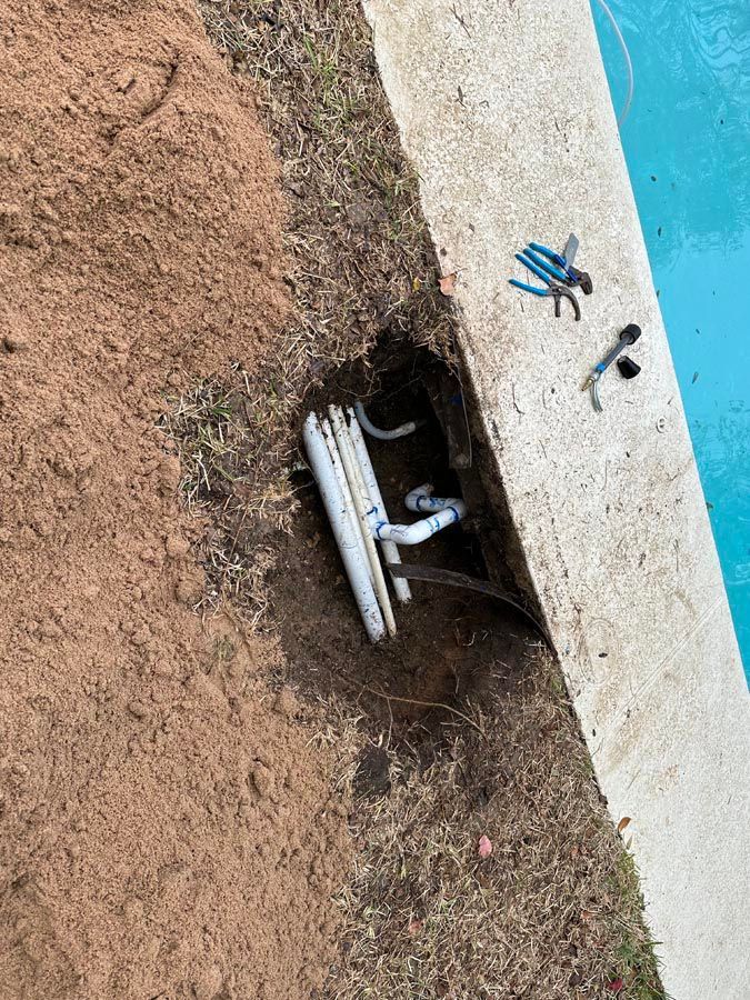 Hole in the ground next to a pool revealing plumbing. Blue pool water, white pipes and tools are visible.
