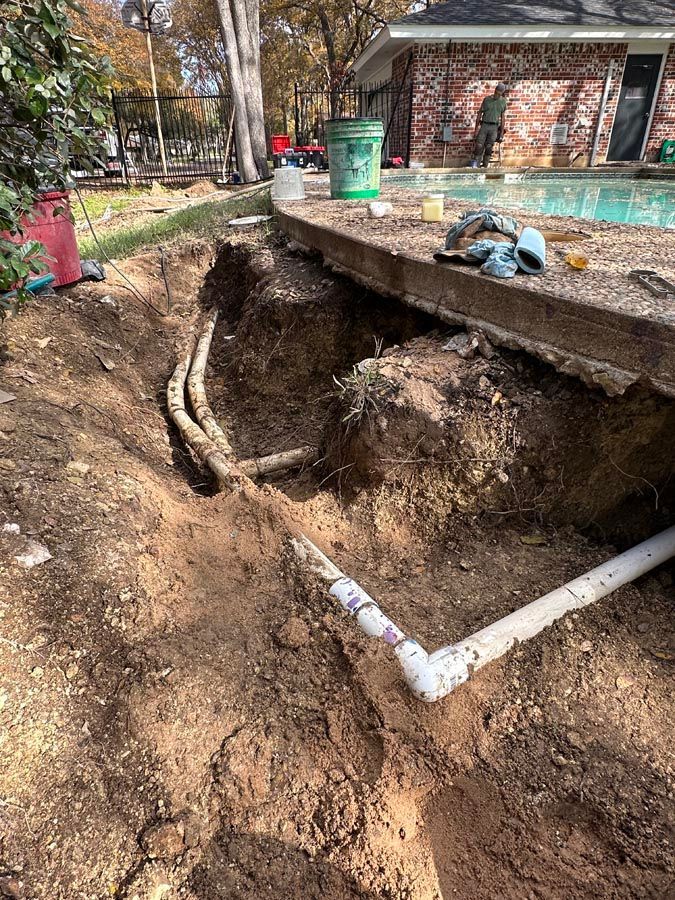 Excavated area near a pool with visible PVC pipes and other plumbing. Dirt and construction materials are present.