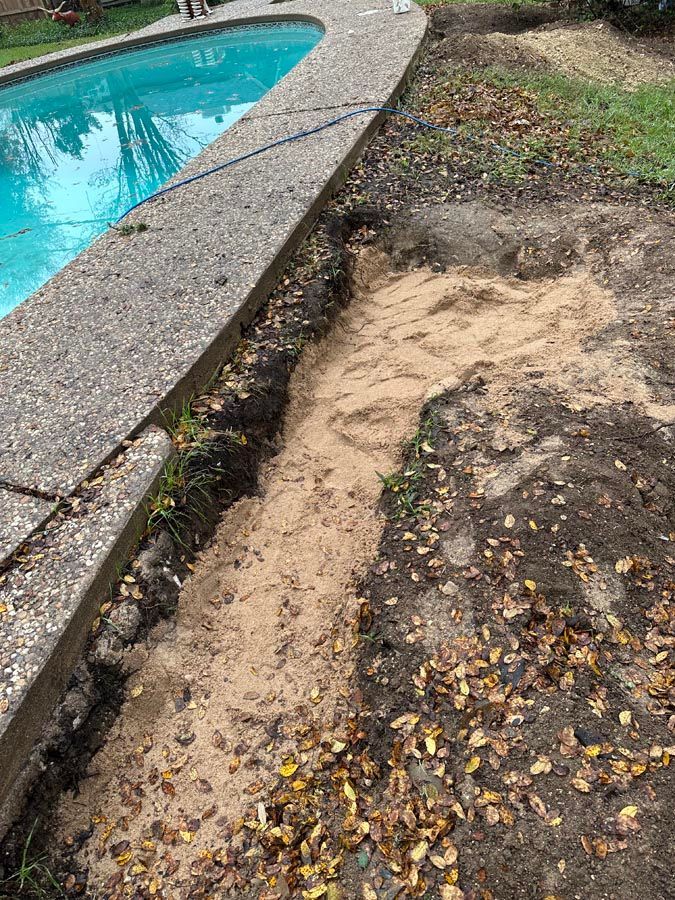 A trench filled with sand runs along a concrete pool edge, dirt and leaves nearby.
