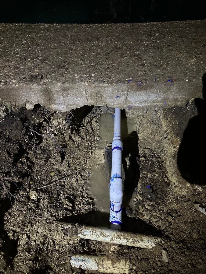 A PVC pipe connection exposed in a trench filled with water; set against concrete and dirt.