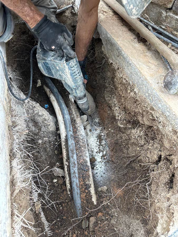 Person using a jackhammer to break concrete around pipes in a trench.