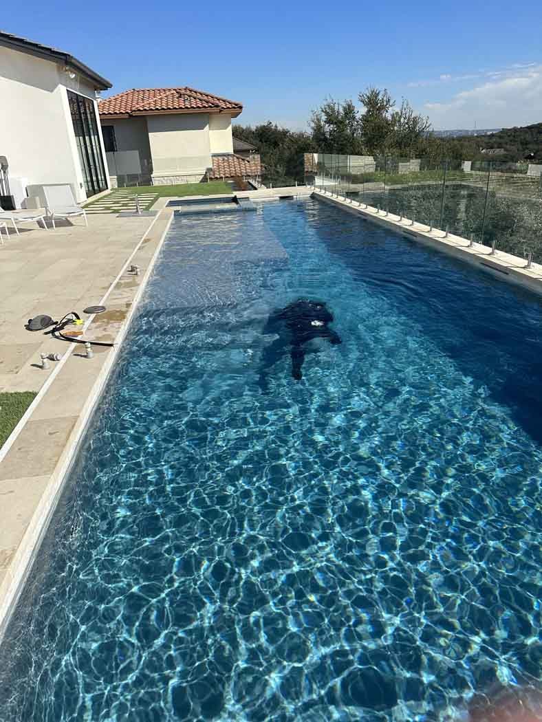 Person swimming in a long, blue outdoor pool with a house and clear sky in the background.