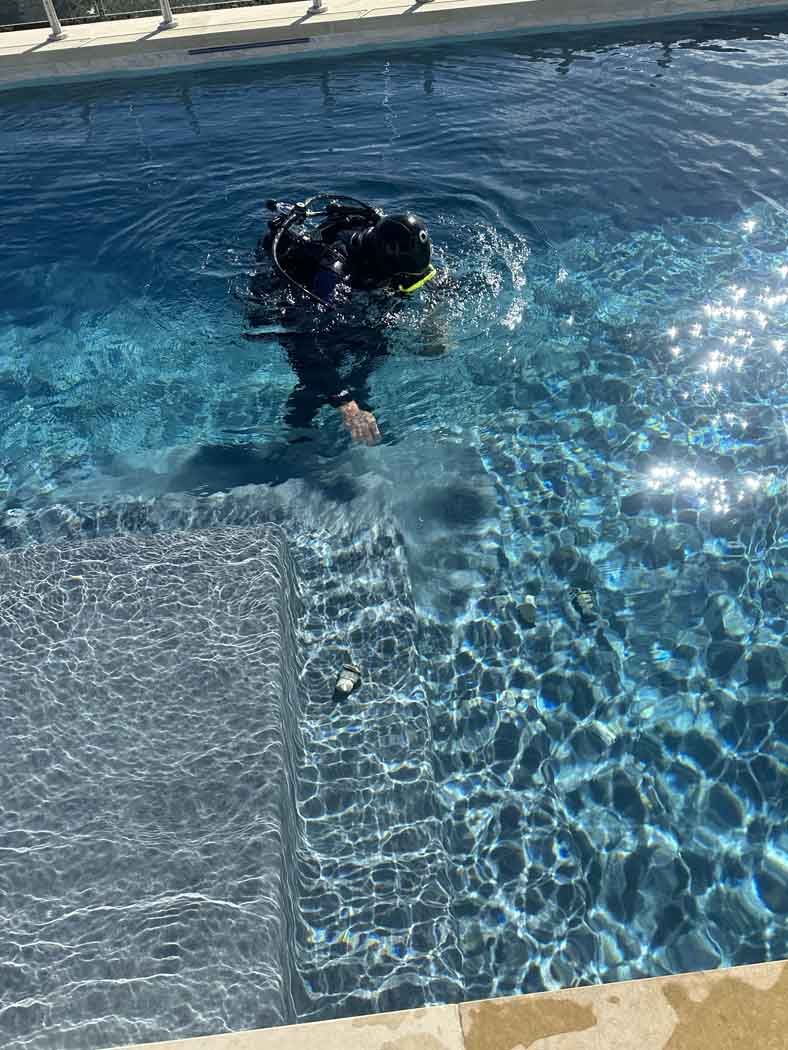 Person wearing scuba gear in a blue pool, near steps. Sunlight reflects on the water's surface.