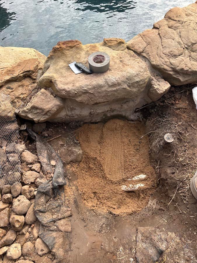 Sand and rocks surround an excavation area near water; duct tape and a trowel sit on a boulder.