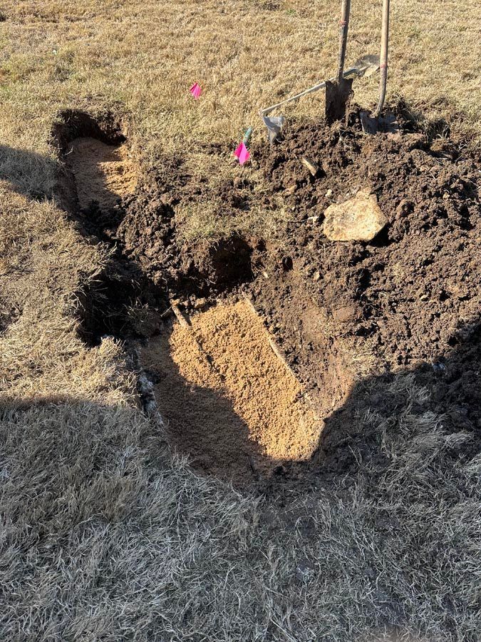 Hole in the ground with upturned soil. Shovel nearby with pink flags and dry grass in the background.