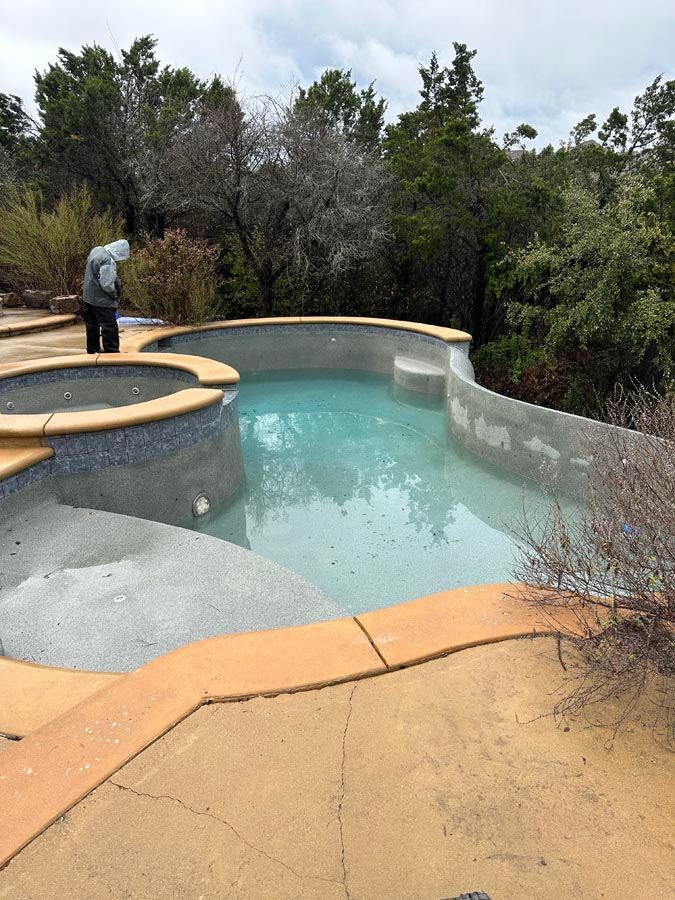Pool partially filled with water, surrounded by tan concrete and natural foliage. Person near edge.