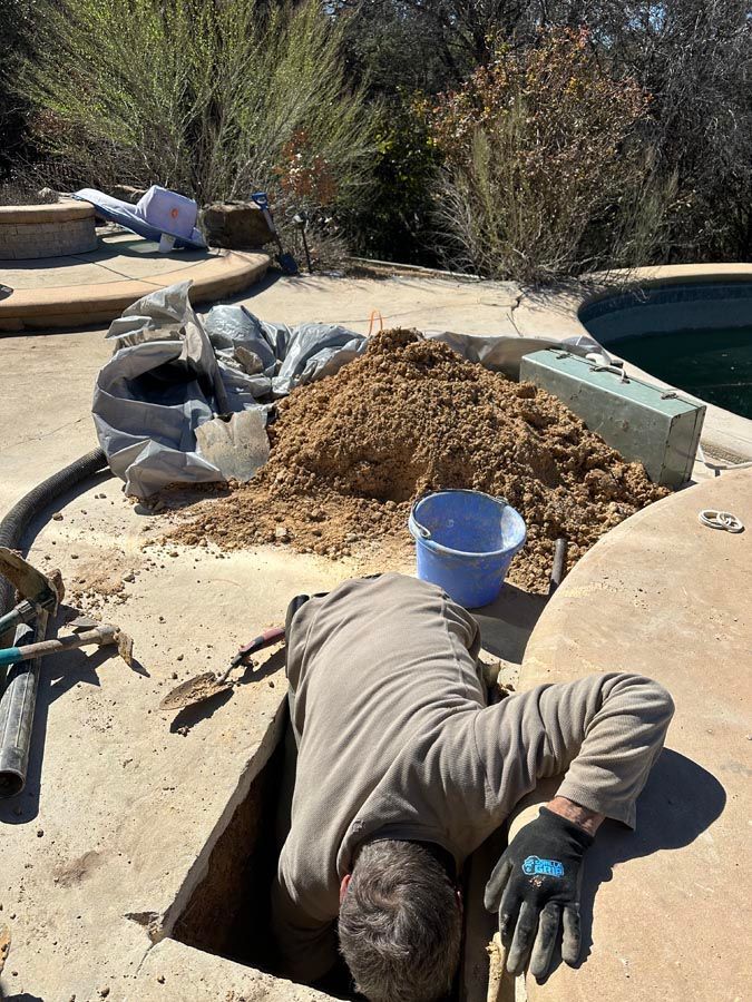 Person working inside a concrete hole near a swimming pool. Sand and debris are piled nearby. Sunny outdoor setting.