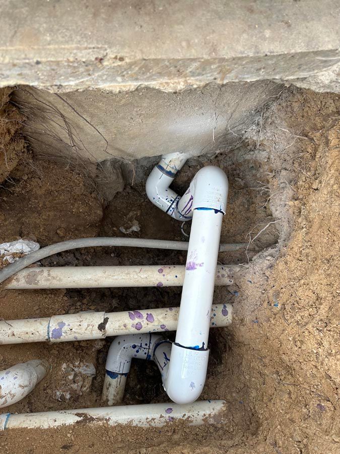 White PVC pipes in a dirt trench, likely plumbing repair. Concrete above.