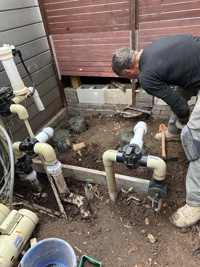 Man working on pool plumbing in a dirt area with pipes and a wooden fence.
