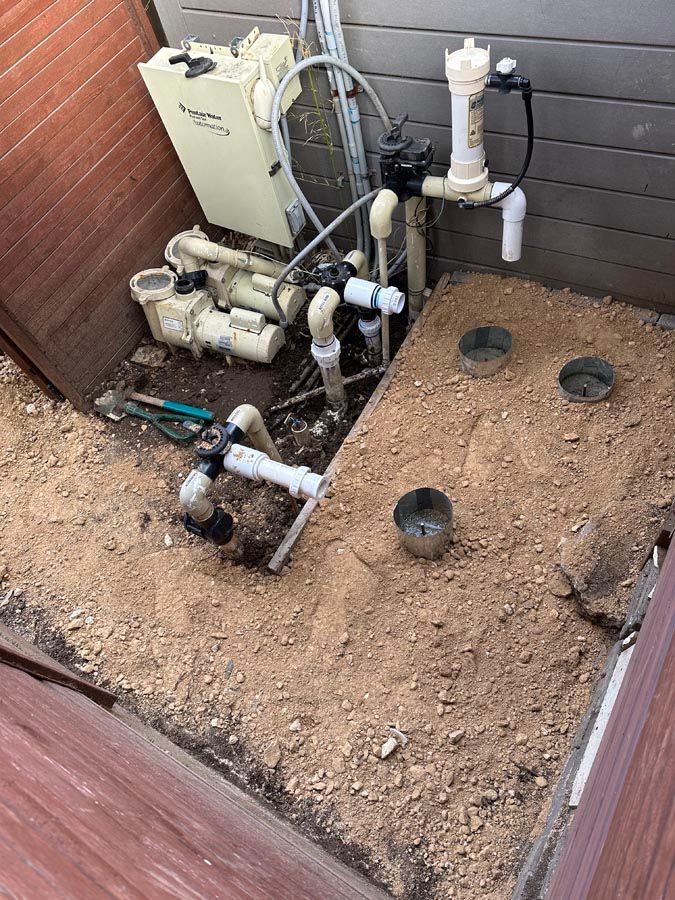 Pool equipment in dirt pit, including pump, filter, and chlorinator, near a wooden structure.