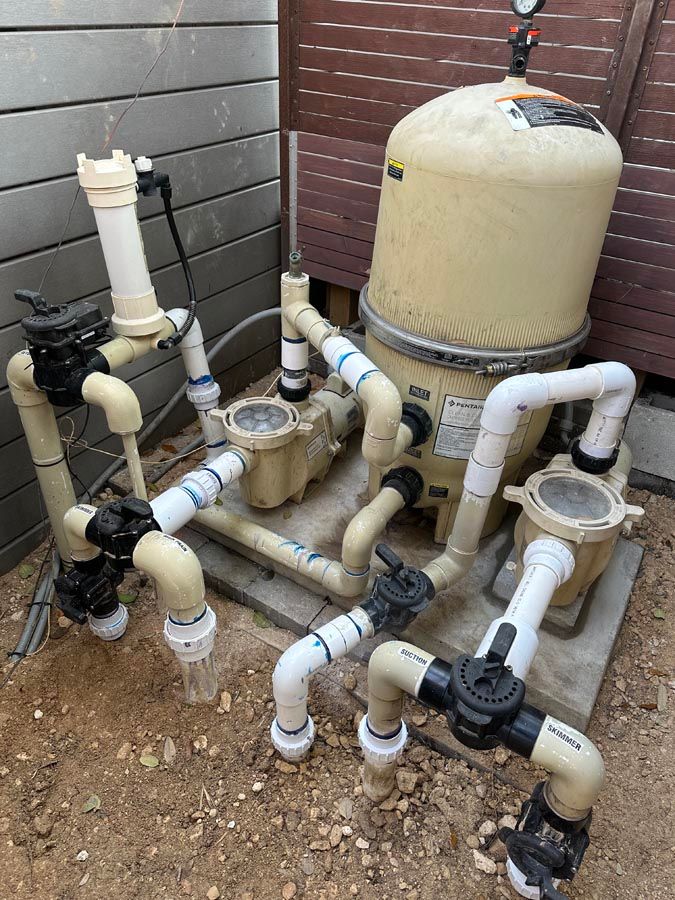Pool equipment: Beige filter, pumps, and pipes with valves on gravel surface, near a wooden fence.