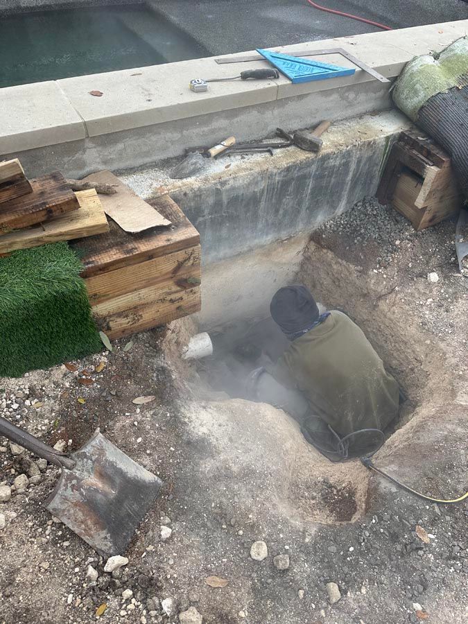 Person in a hole near a pool, digging. Dust is visible. Tools, wood, and a pool surround are also present.