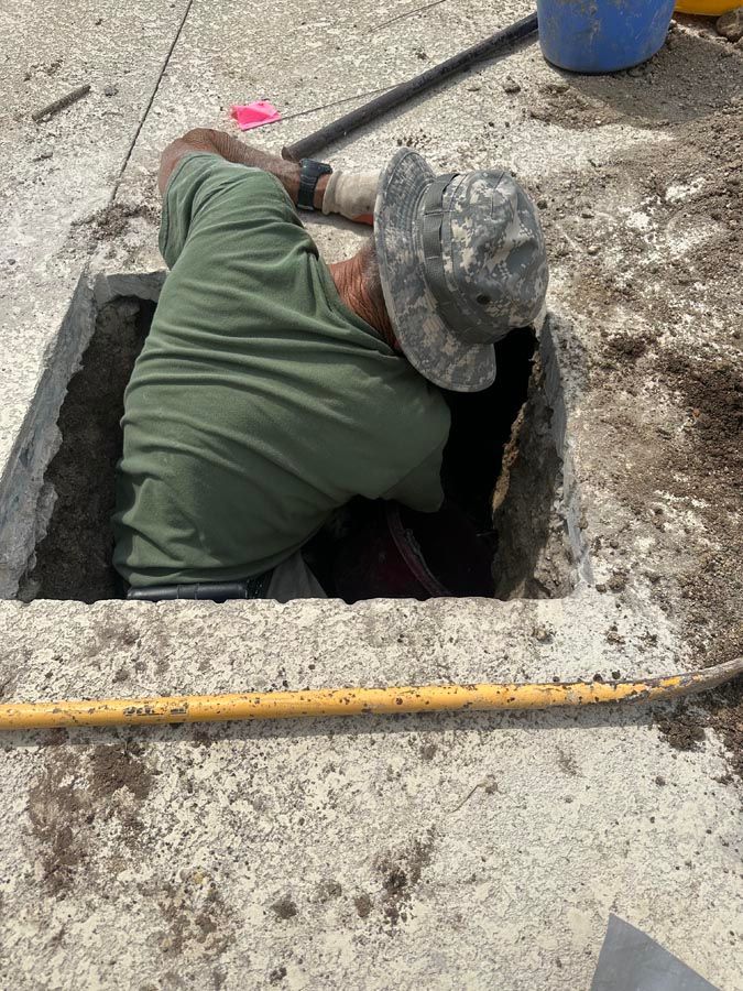 Person leaning into a rectangular hole in concrete, wearing a green shirt and camouflage hat; working on something underground.