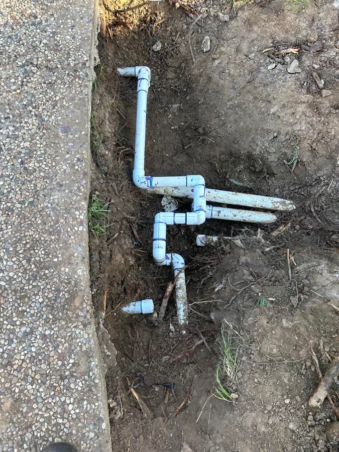Blue PVC pipes in a dirt trench next to a concrete walkway, for an irrigation system.