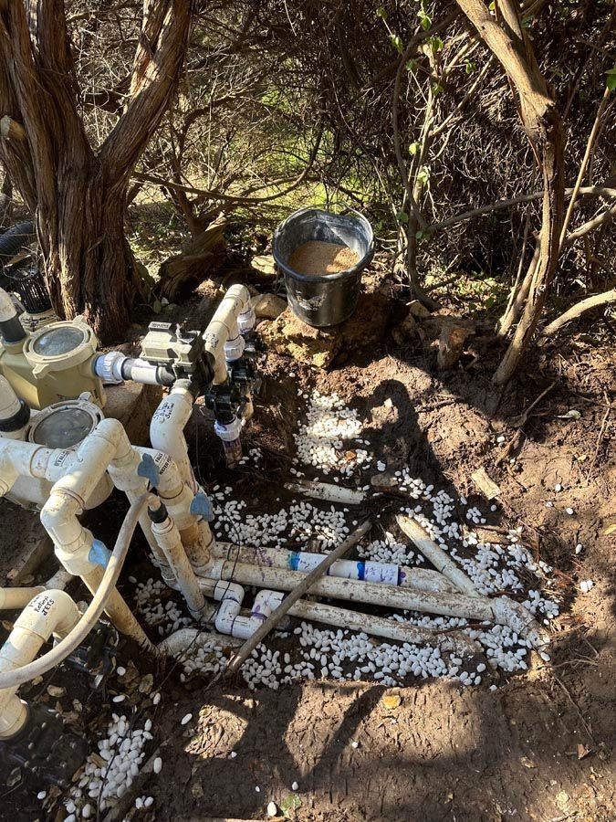 Pool plumbing: white pipes, valves, and filter components disassembled outdoors in dirt and gravel, with a bucket.