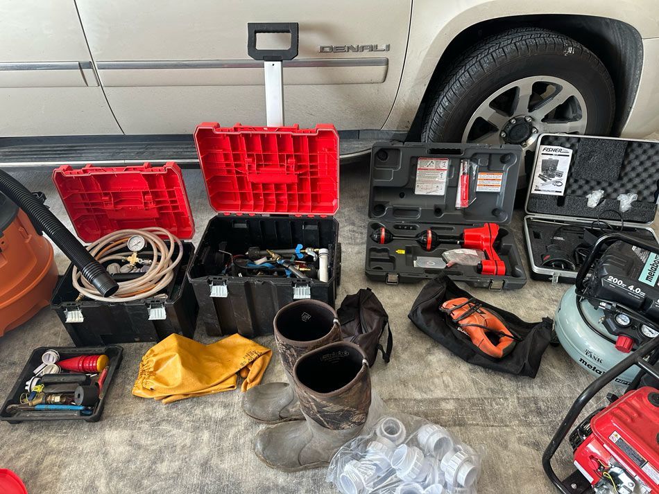 Tools and equipment, including drills, cases, a vacuum, boots, and pipes, are arranged in front of a white vehicle.