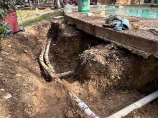 Trench dug near a pool, exposing pipes. Dirt, pool deck, and tools visible.