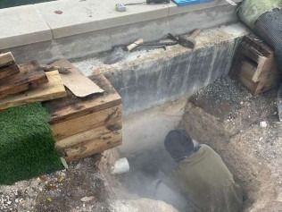 Man digging in a trench near a concrete wall. Dust is visible, tools and wood are nearby.