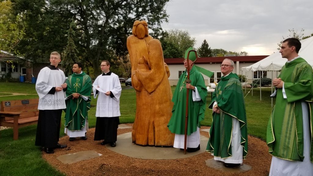 Clergy in green and white robes stand around a wooden statue on a lawn during an outdoor ceremony