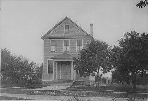 Black-and-white photo of a two-story house with a porch, surrounded by trees and lawn.