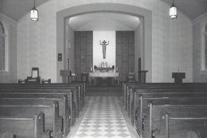 Black-and-white view of a small church interior with pews, tiled aisle, and altar at the far end.